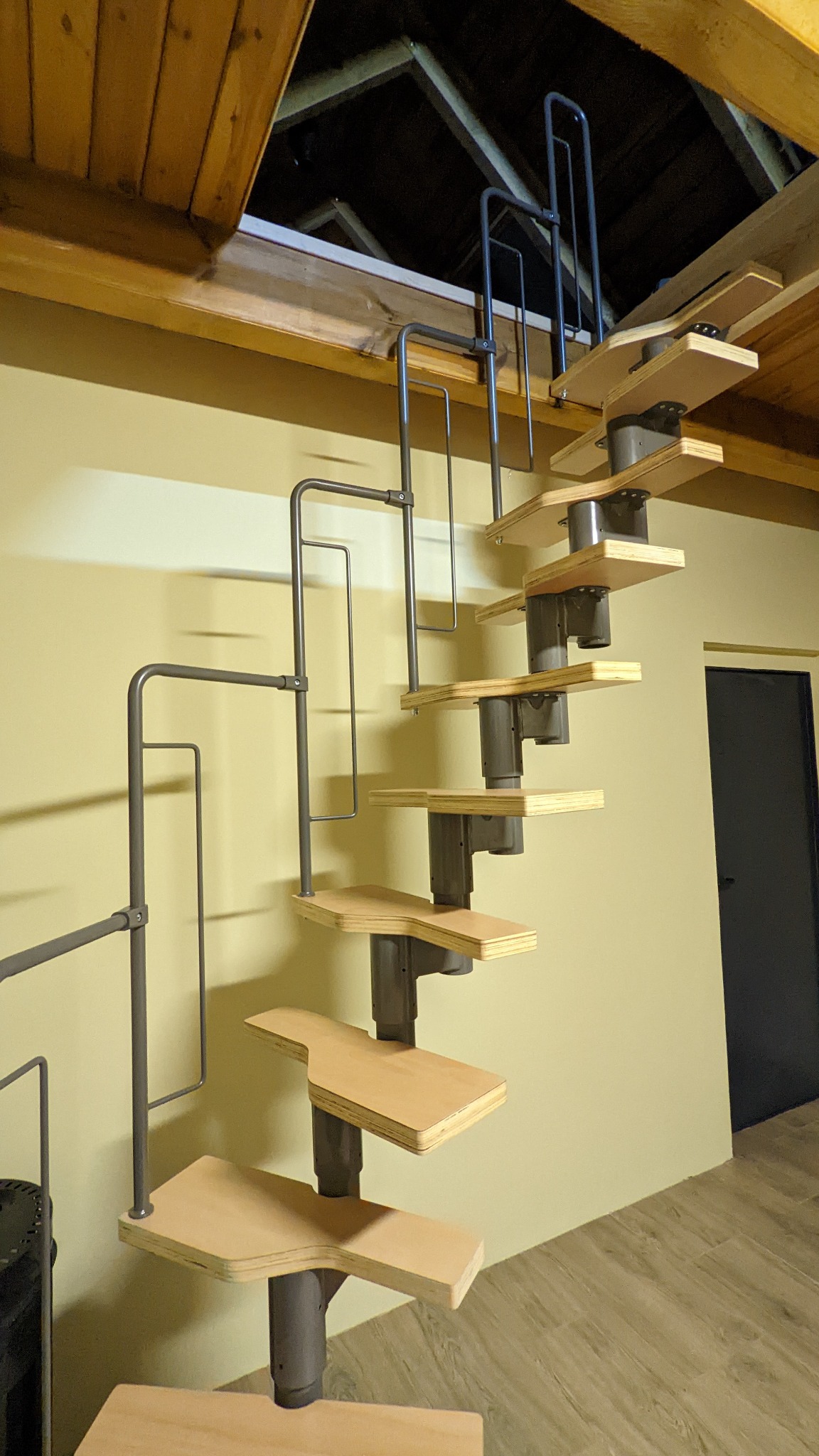 Spiralne schody strychowe z drewnianymi stopniami i metalową konstrukcją, prowadzące do otworu w drewnianym suficie poddasza.