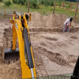 Żółta koparka JCB wykopuje rów pod fundament, pracownik wyrównuje ziemię łopatą, widok z kabiny operatora, w tle krajobraz z drzewami i pochmurnym niebem.