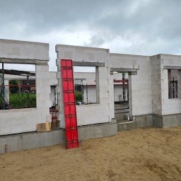 AS Sławomir Krajewski - Budowa parterowego domu z białych bloczków komórkowych, z widocznymi otworami okiennymi i czerwonymi szalunkami, na piaszczystym placu budowy pod zachmurzonym niebem.