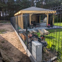 AS Sławomir Krajewski - Ciemne ogrodzenie panelowe na betonowej podmurówce, oddzielające piaszczysty teren od drewnianej altany z szarym dachem, brukowanego patio i zielonego trawnika.