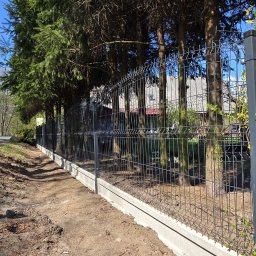 AS Sławomir Krajewski - Nowo zainstalowane ciemnoszare ogrodzenie panelowe z betonową podmurówką, biegnące wzdłuż ścieżki obok rzędu wysokich drzew iglastych. W tle widoczny fragment domu.
