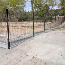 AS Sławomir Krajewski - Czarne ogrodzenie panelowe z betonowymi podmurówkami, biegnące wzdłuż pochyłego terenu obok utwardzonej ścieżki. W tle drzewa i biały van z otwartymi drzwiami, przy którym stoi osoba.