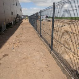 AS Sławomir Krajewski - Nowe grafitowe ogrodzenie panelowe z betonową podmurówką, biegnące wzdłuż nowoczesnej hali przemysłowej. Piaszczysty teren budowy w słoneczny dzień.
