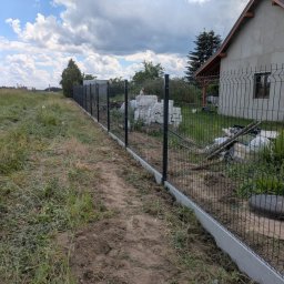 AS Sławomir Krajewski - Ukończony montaż ciemnoszarego ogrodzenia panelowego z betonową podmurówką, biegnącego wzdłuż ścieżki. W tle dom i stos bloczków budowlanych.