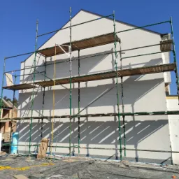 Budynek w trakcie prac elewacyjnych, widoczne rusztowanie, ocieplenie styropianem oraz jasna, strukturalna nowa elewacja. W tle nieukończony budynek z cegły.