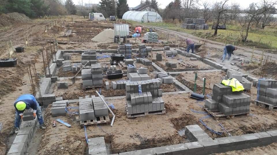 Budowa fundamentów domu z bloczków betonowych: pracownicy układają bloczki na przygotowanym fundamencie, widoczne zbrojenia, palety z bloczkami, ziemia i budynki w tle.