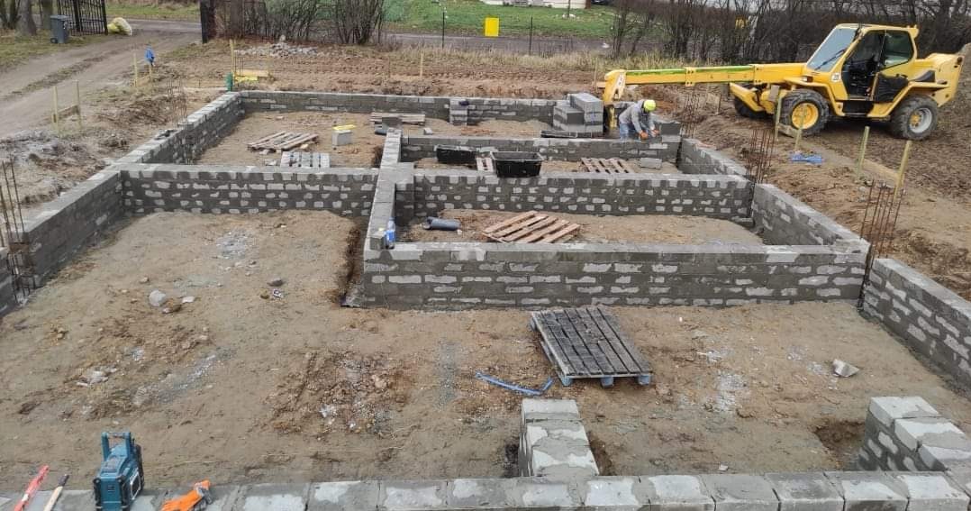 Wykopane i częściowo wymurowane fundamenty pod budynek z widocznym pracownikiem i żółtą maszyną budowlaną w tle.