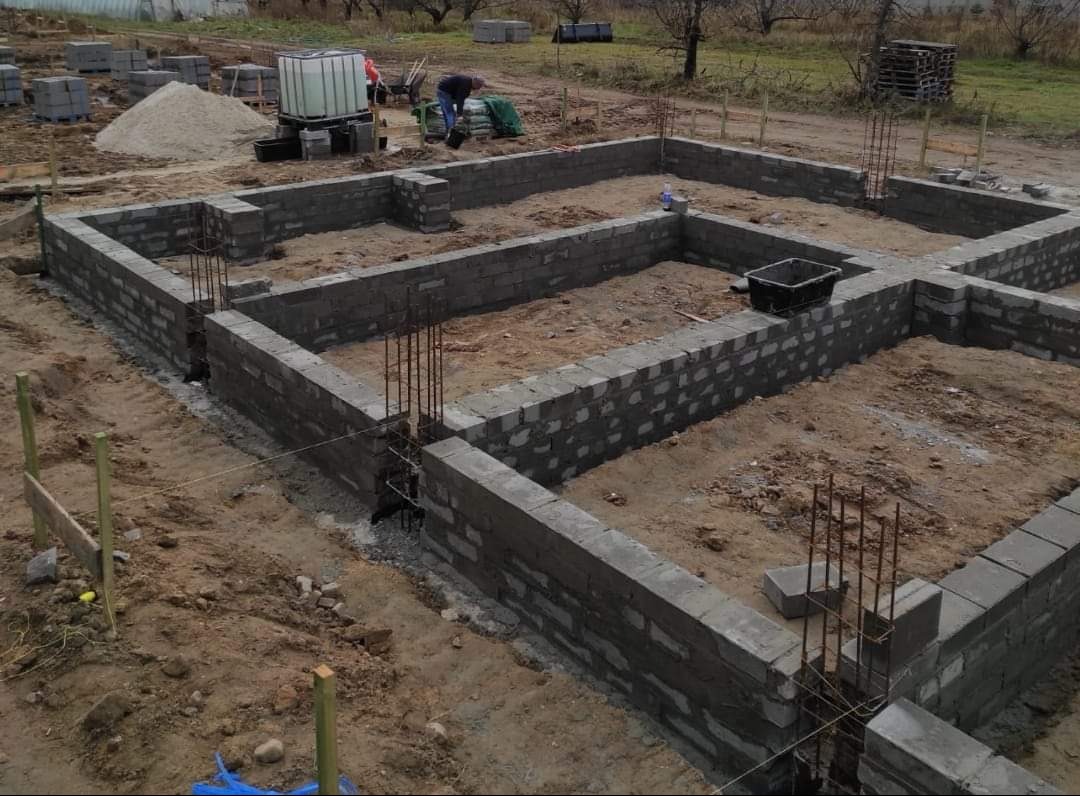 Wznoszenie fundamentów z bloczków betonowych, widoczne wystające pręty zbrojeniowe, zaplecze budowy z paletami i zbiornikiem na wodę w tle.