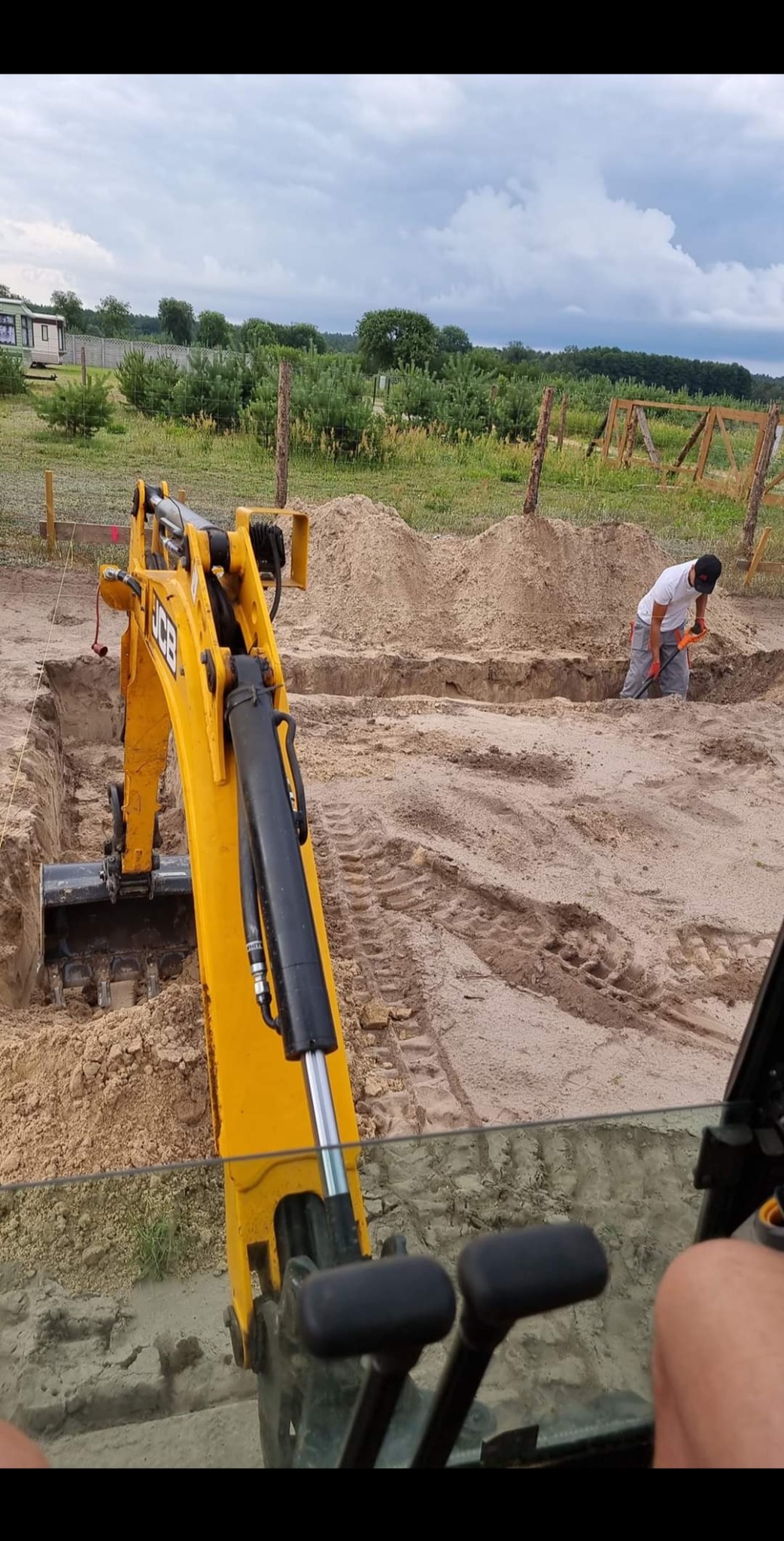 Żółta koparka JCB wykopuje rów pod fundament, pracownik wyrównuje ziemię łopatą, widok z kabiny operatora, w tle krajobraz z drzewami i pochmurnym niebem.