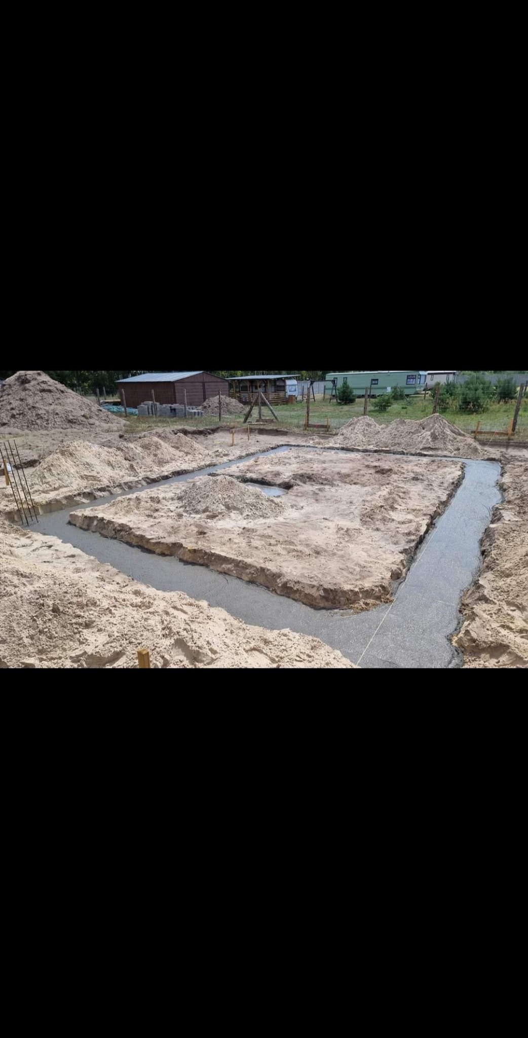 Wylane fundamenty pod budynek na piaszczystym terenie, widoczne zbrojenia i drewniane szalunki w tle.