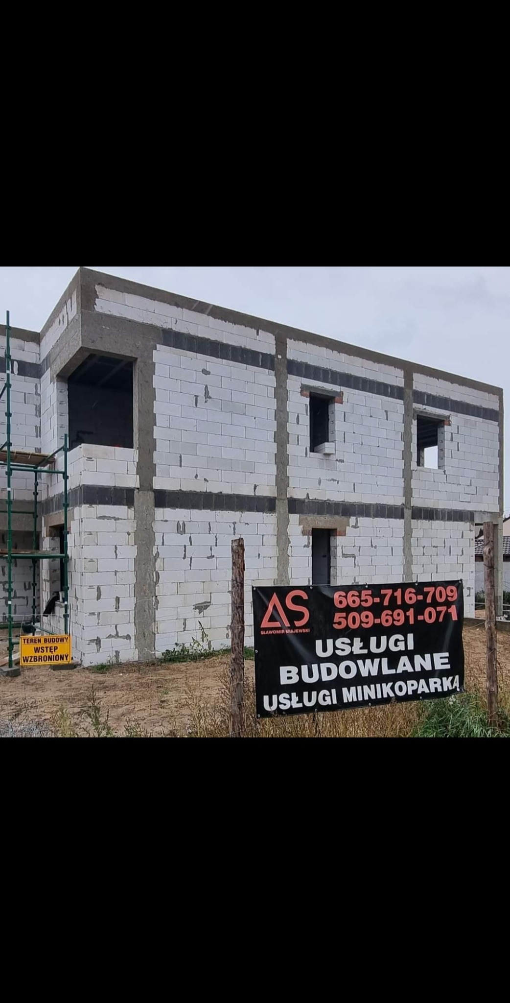 Dwukondygnacyjny dom murowany w trakcie budowy z białych bloczków, z widocznymi otworami okiennymi i drzwiowymi, częściowo osadzony w surowym betonie, z rusztowaniami z boku i tablicą informacyjną...