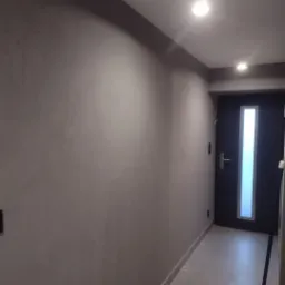 Korytarz po nałożeniu szarej, dekoracyjnej gładzi z widoczną fakturą, oświetlony punktowo, z ciemnymi drzwiami z długim, pionowym przeszkleniem.