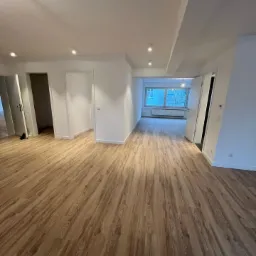 Jasne panele podłogowe z widoczną strukturą drewna w wyremontowanym, pustym pokoju z białymi ścianami i sufitami oraz zamontowanym oświetleniem punktowym.