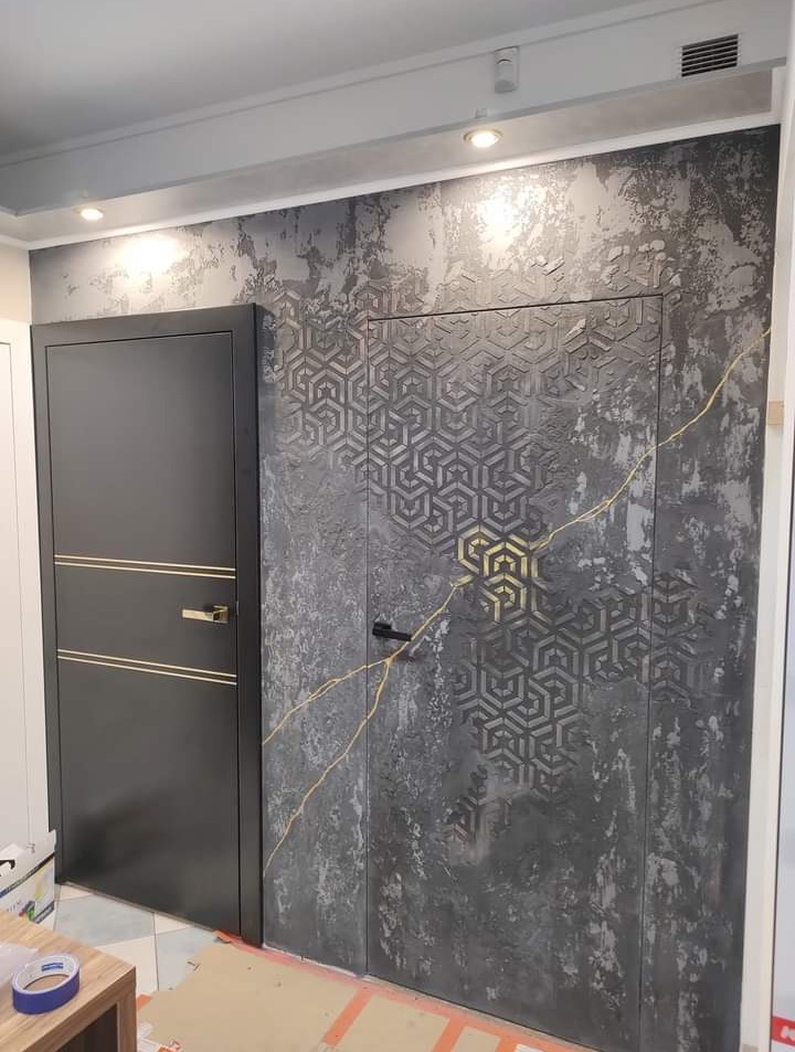 Dwie nowoczesne, ciemne drzwi wewnętrzne: Jedna z prostymi złotymi paskami, druga z geometrycznym wzorem na tle ściany imitującej surowy beton z efektem popękanej złotej żyły.