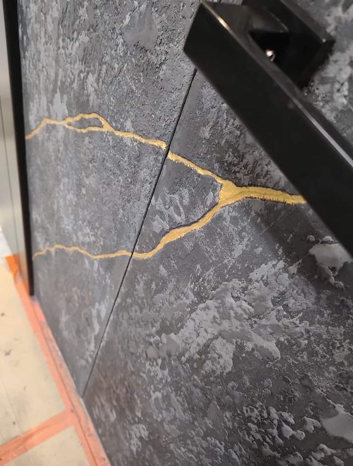 Dekoracyjna ściana z szarą, chropowatą fakturą, zdobiona nieregularnymi, złotymi żyłkami, widoczna czarna klamka i fragment zabezpieczonej podłogi.