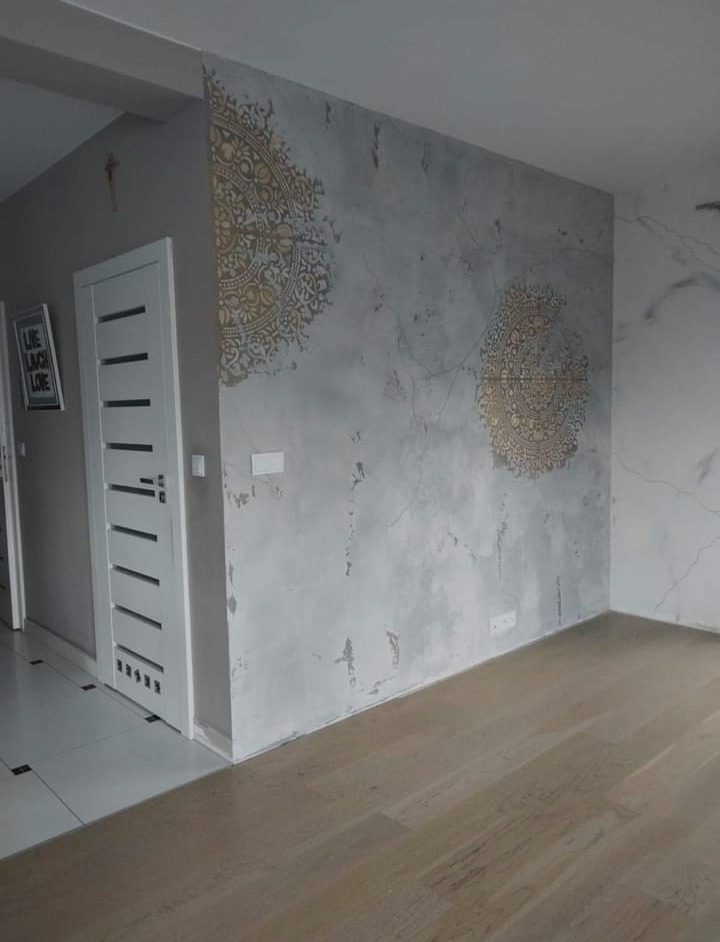 Ściana w pokoju z szarą dekoracyjną fakturą i dwoma złotymi ornamentami w stylu mandali, jasna podłoga z paneli, białe drzwi z poziomymi frezowaniami.