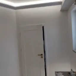 Nowoczesna, minimalistyczna łazienka z białymi ścianami, podświetlanym sufitem i częściowo otwartymi białymi drzwiami; widoczna umywalka z marmurowym blatem i akcesoriami.