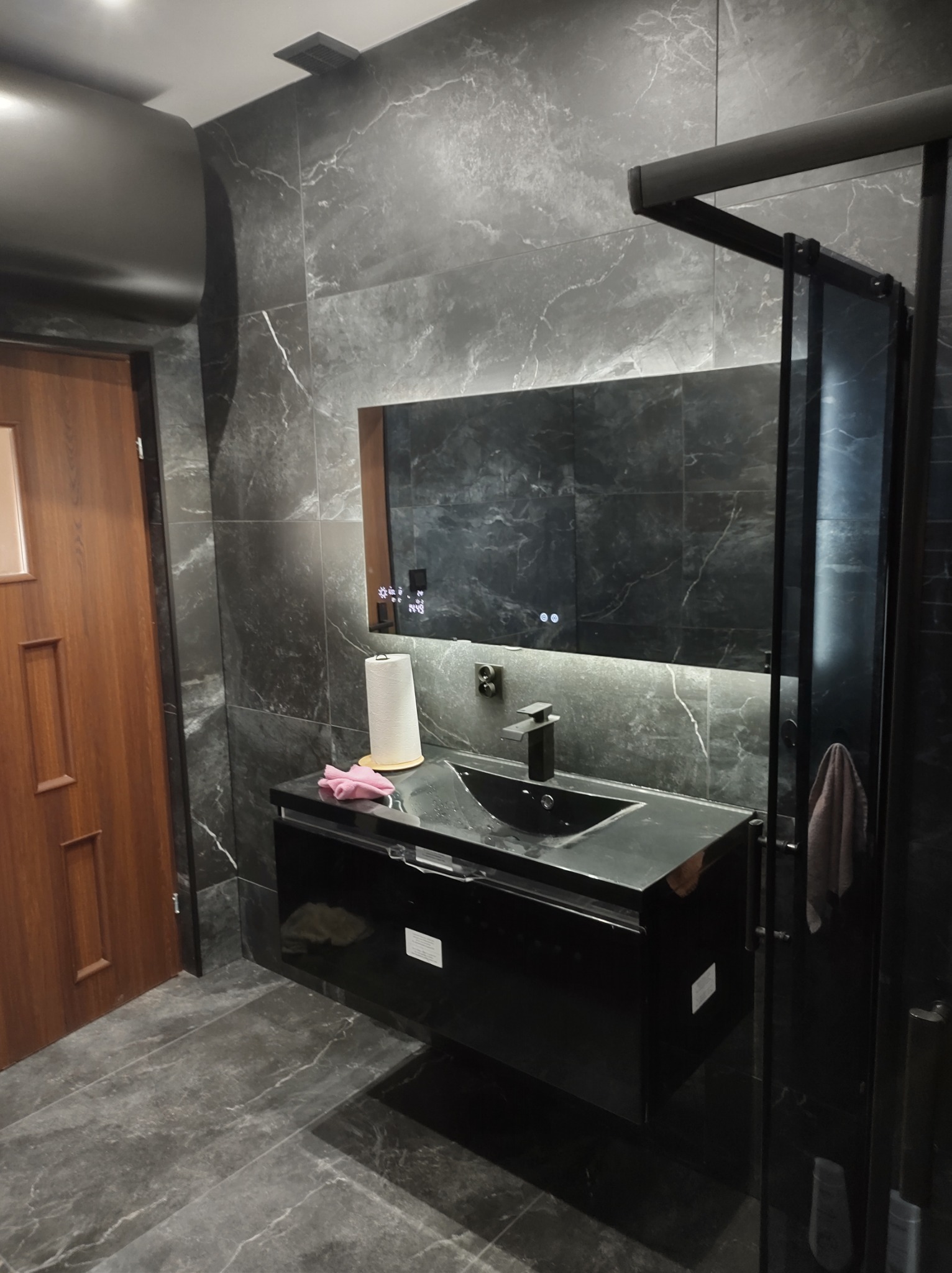 Nowoczesna łazienka z czarnymi płytkami imitującymi marmur, czarną szafką pod umywalką z podświetlanym lustrem i czarną armaturą oraz kabiną prysznicową z czarnymi profilami.