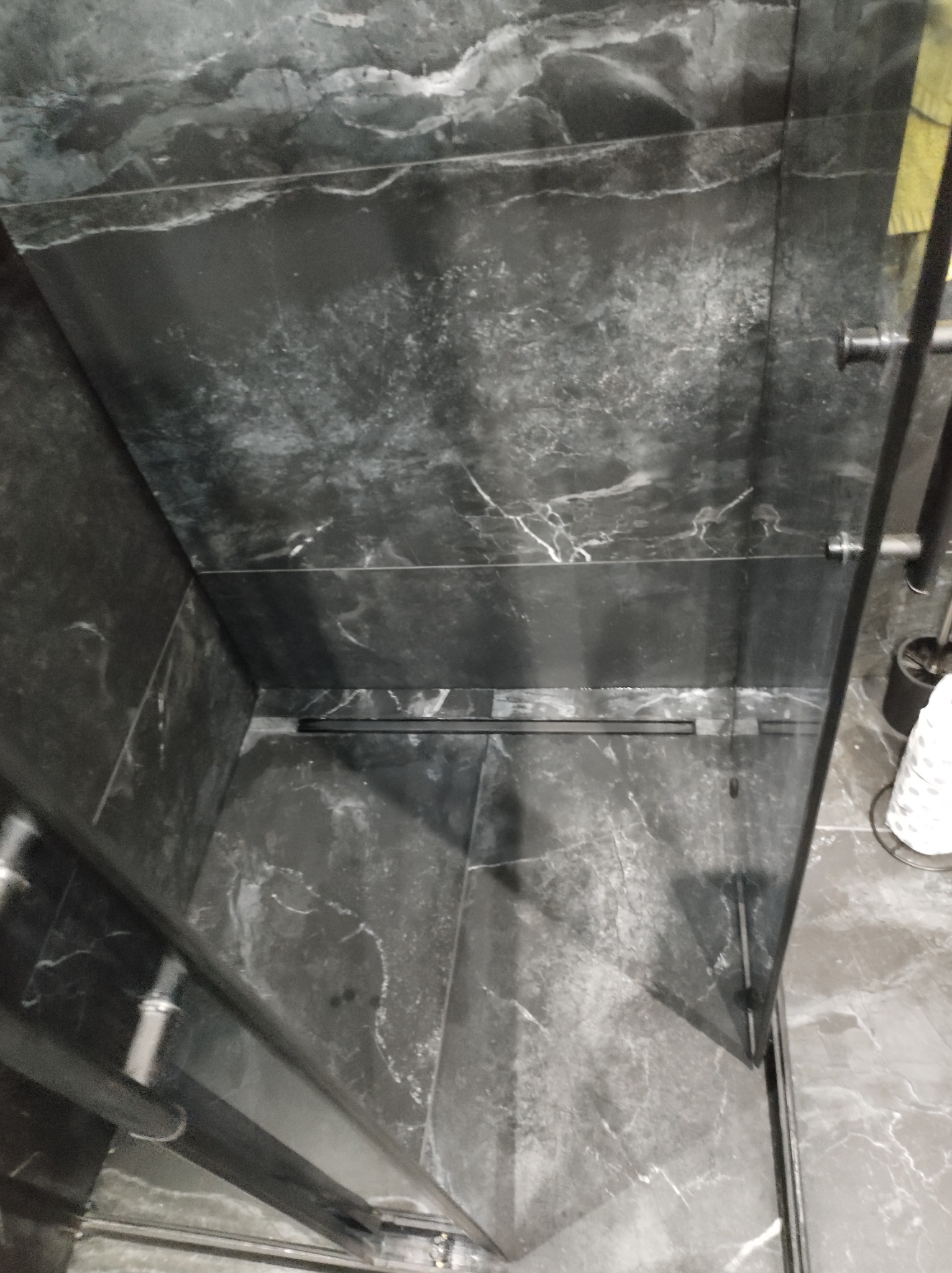 Nowoczesna kabina prysznicowa z czarnymi, marmurkowymi płytkami, widoczny odpływ liniowy w podłodze i szklane drzwi z czarnymi profilami.