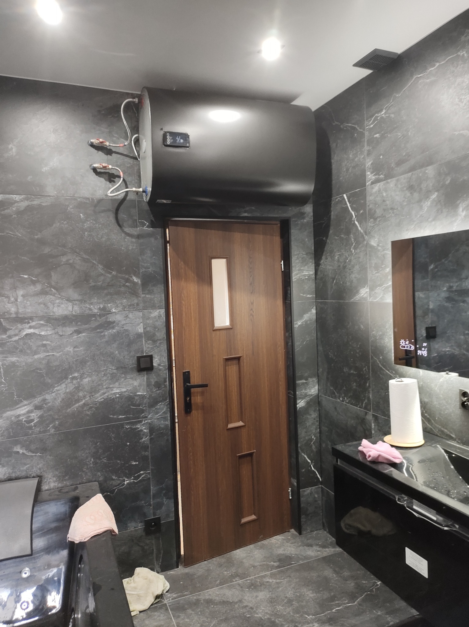 Ciemnoszara łazienka z marmurowymi płytkami, wiszący bojler, drewniane drzwi z szybką, czarna szafka umywalkowa i cyfrowym lustrem.