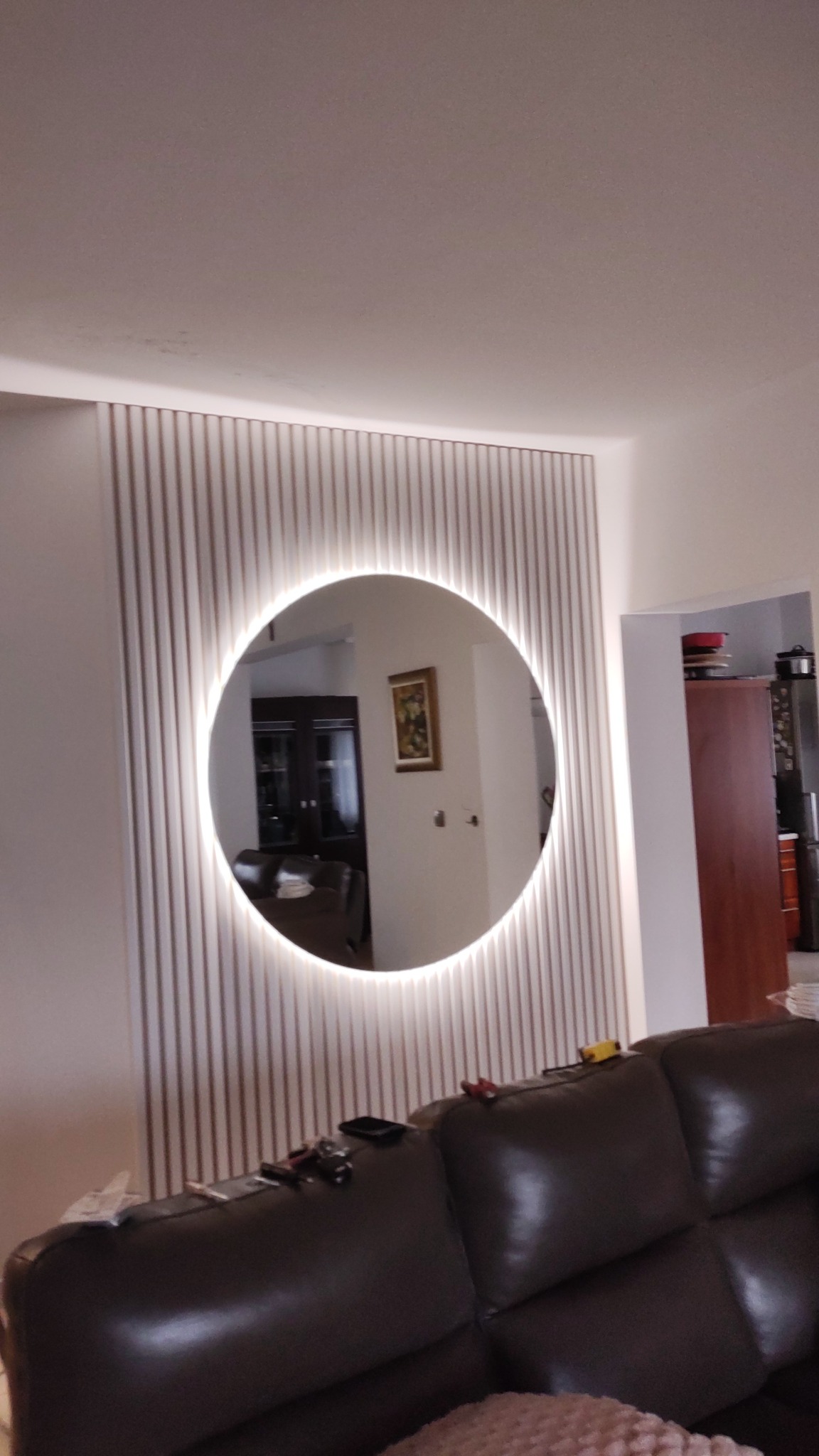 Okrągłe lustro z podświetleniem LED, zamontowane na ścianie z dekoracyjnych paneli pionowych, odbijające wnętrze pokoju z obrazem i meblami.