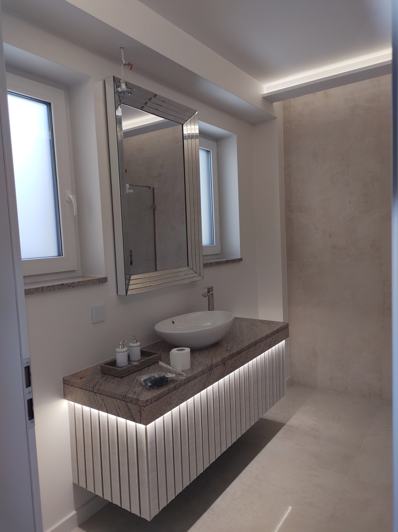 Nowoczesna łazienka z podświetlaną szafką pod umywalką z blatem z kamienia, lustro w ozdobnej ramie, minimalistyczna armatura i beżowe płytki.