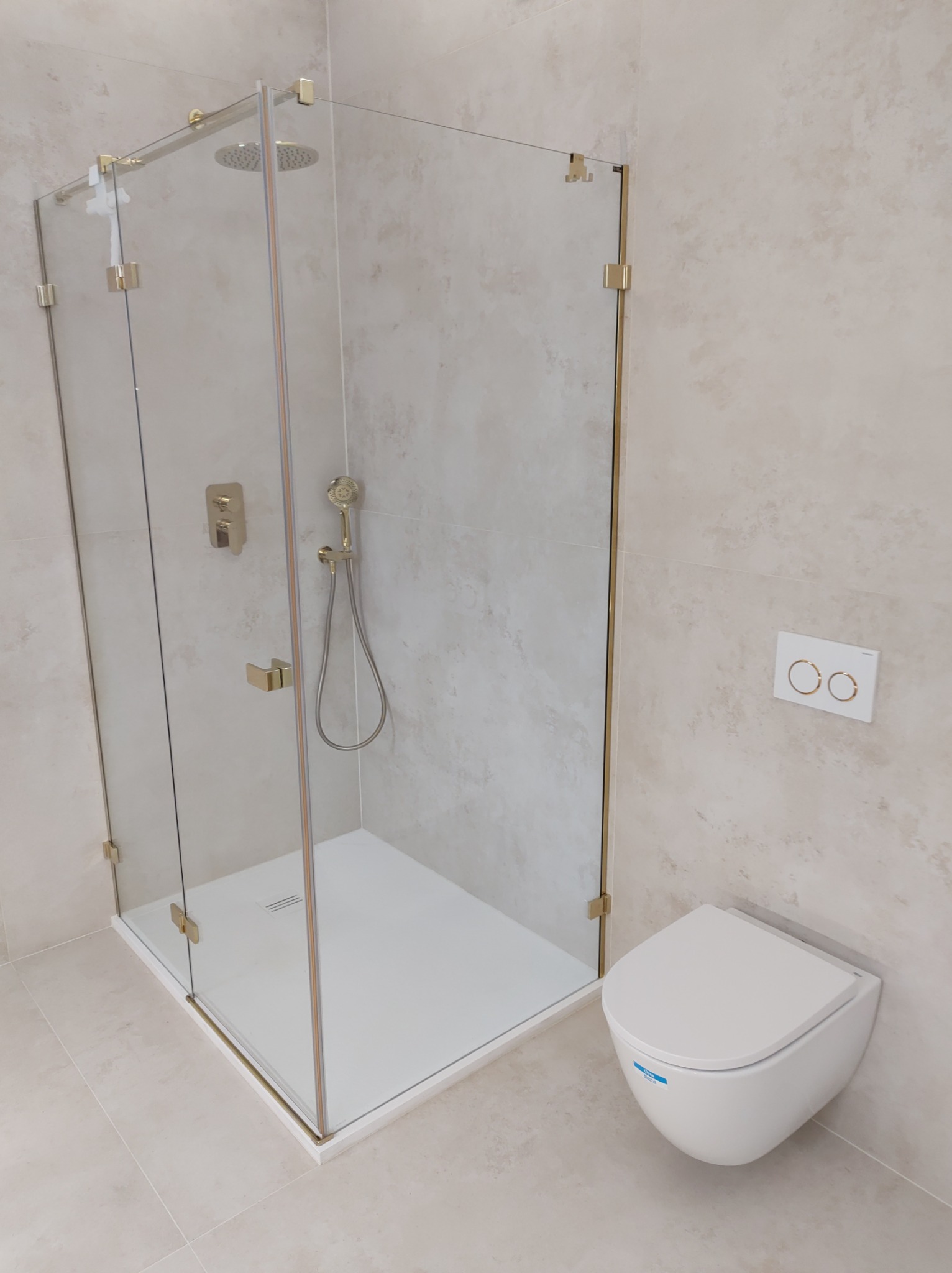 Nowoczesna kabina prysznicowa z elementami w kolorze złotym, dopasowana do wiszącej miski WC w łazience wykończonej jasnymi płytkami.