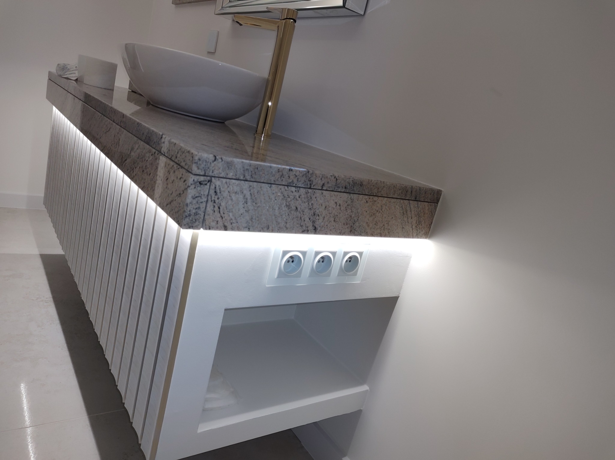 Nowoczesna łazienka z blatem z szarego kamienia, nablatową umywalką, złotą baterią i podświetlanymi listwami pionowymi oraz gniazdkami elektrycznymi.