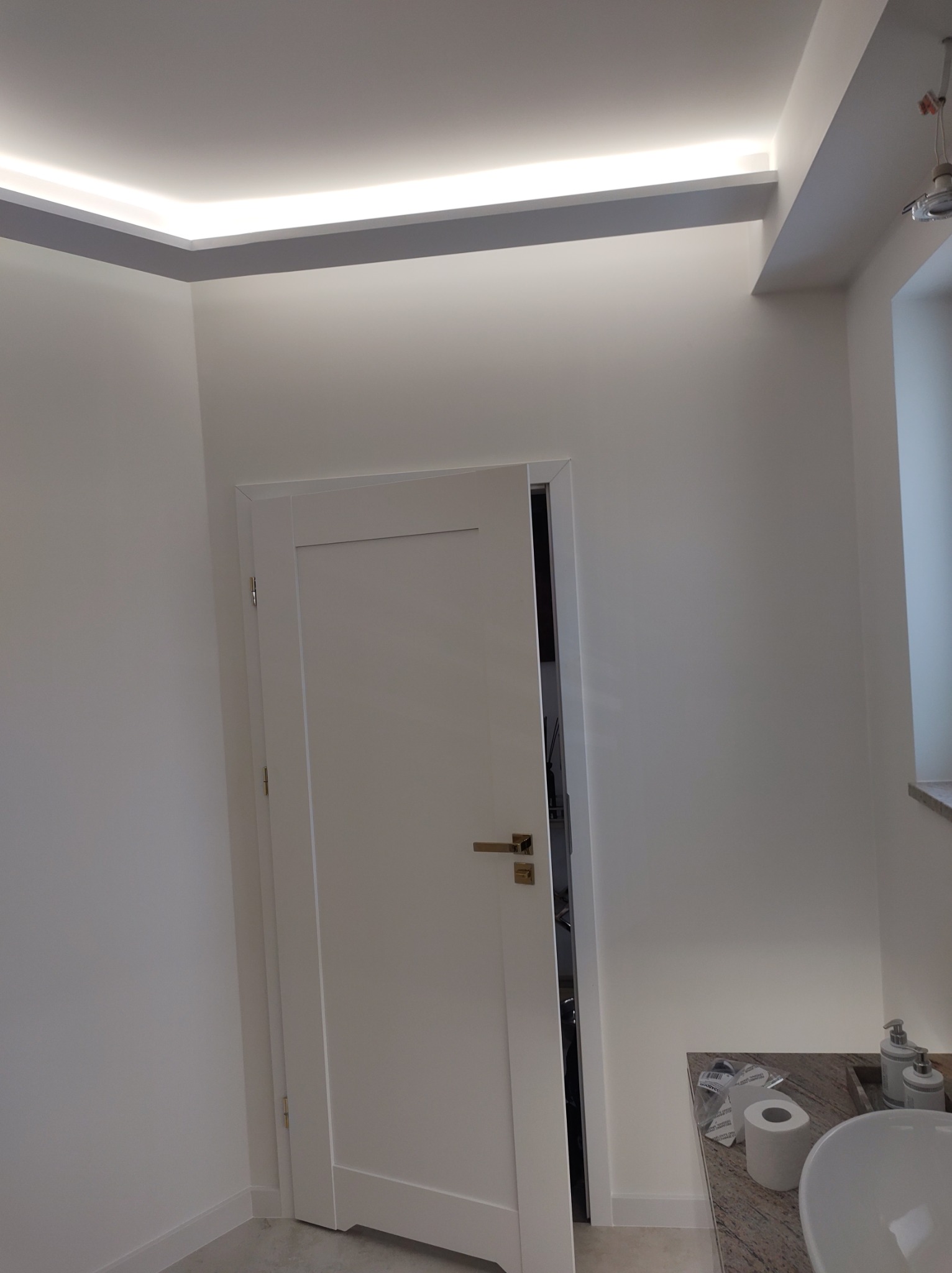 Nowoczesna, minimalistyczna łazienka z białymi ścianami, podświetlanym sufitem i częściowo otwartymi białymi drzwiami; widoczna umywalka z marmurowym blatem i akcesoriami.