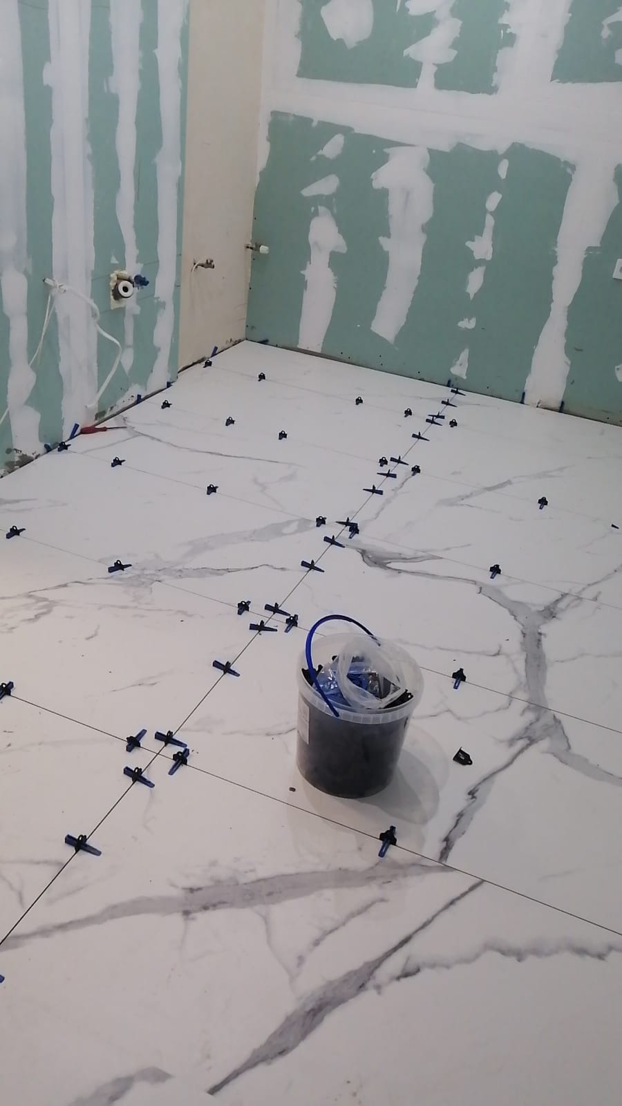 Łazienka w trakcie remontu: białe płytki imitujące marmur na podłodze, krzyżyki dystansowe, ściany przygotowane pod wykończenie, widoczny kubeł z krzyżykami.