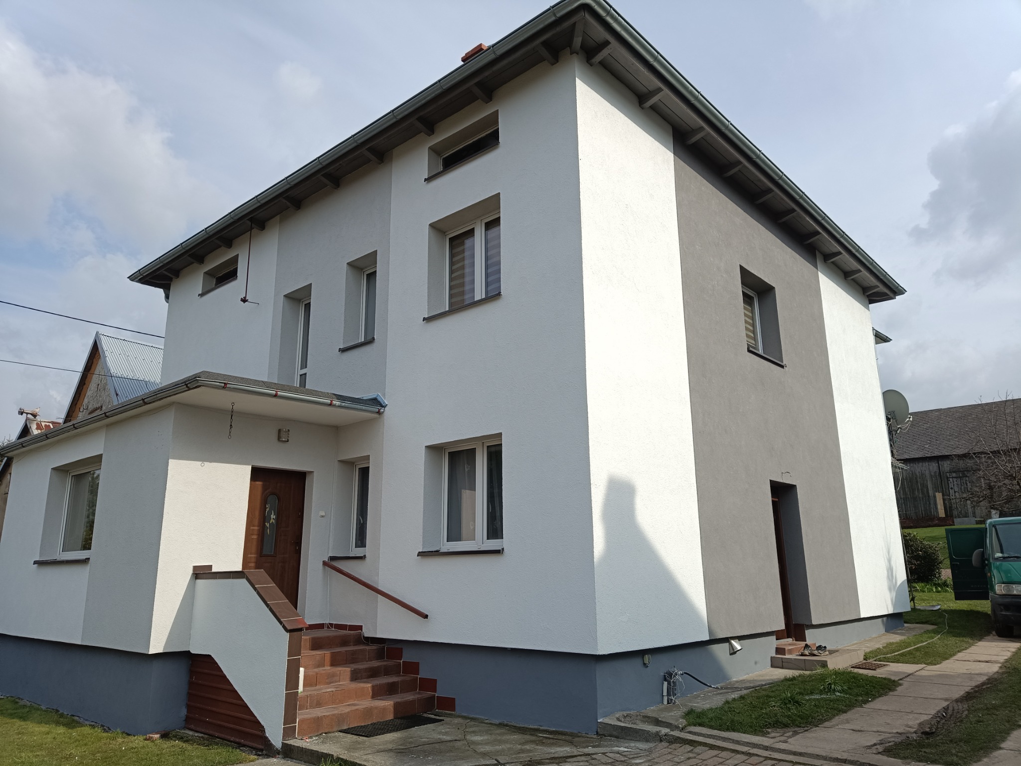 Dwukondygnacyjny dom po ociepleniu ścian zewnętrznych, z częścią elewacji w kolorze białym i szarym, brązowe schody wejściowe i ciemna stolarka okienna.
