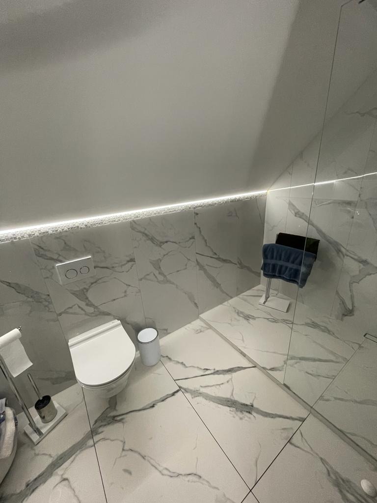 Nowoczesna łazienka na poddaszu z białymi marmurkowymi płytkami, podświetleniem LED i szklaną kabiną prysznicową, widok z góry.