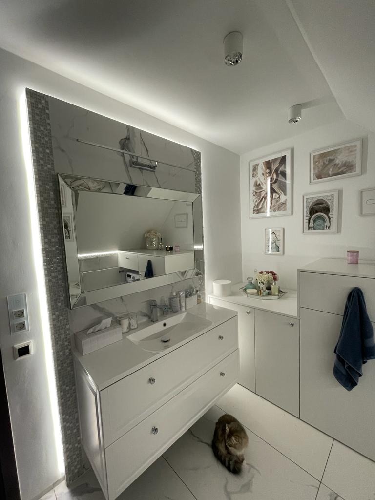 Jasna łazienka w stylu glamour z marmurowymi akcentami, podświetlanym lustrem i białymi meblami. Kot leży na marmurowej podłodze.