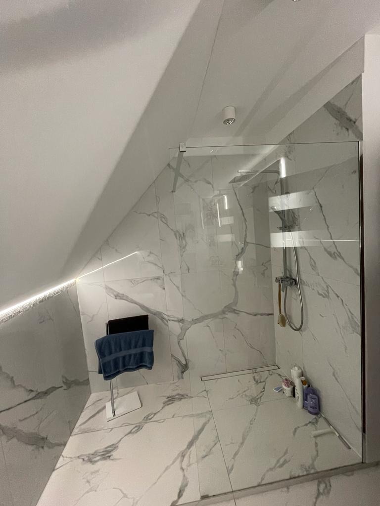 Nowoczesna kabina prysznicowa ze szklaną ścianą w łazience na poddaszu, białe płytki imitujące marmur, chromowana armatura i grzejnik drabinkowy, widoczne kosmetyki na podłodze.