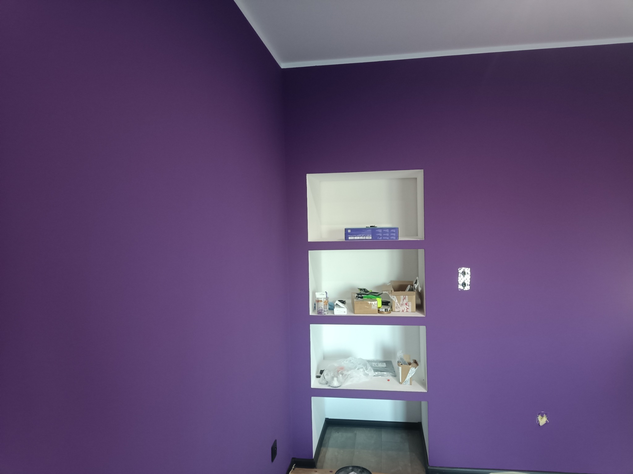 Ściana pomalowana na fioletowo z trzema wnękami, w których znajdują się różne przedmioty, widoczne niedoskonałości w malowaniu i instalacji elektrycznej.