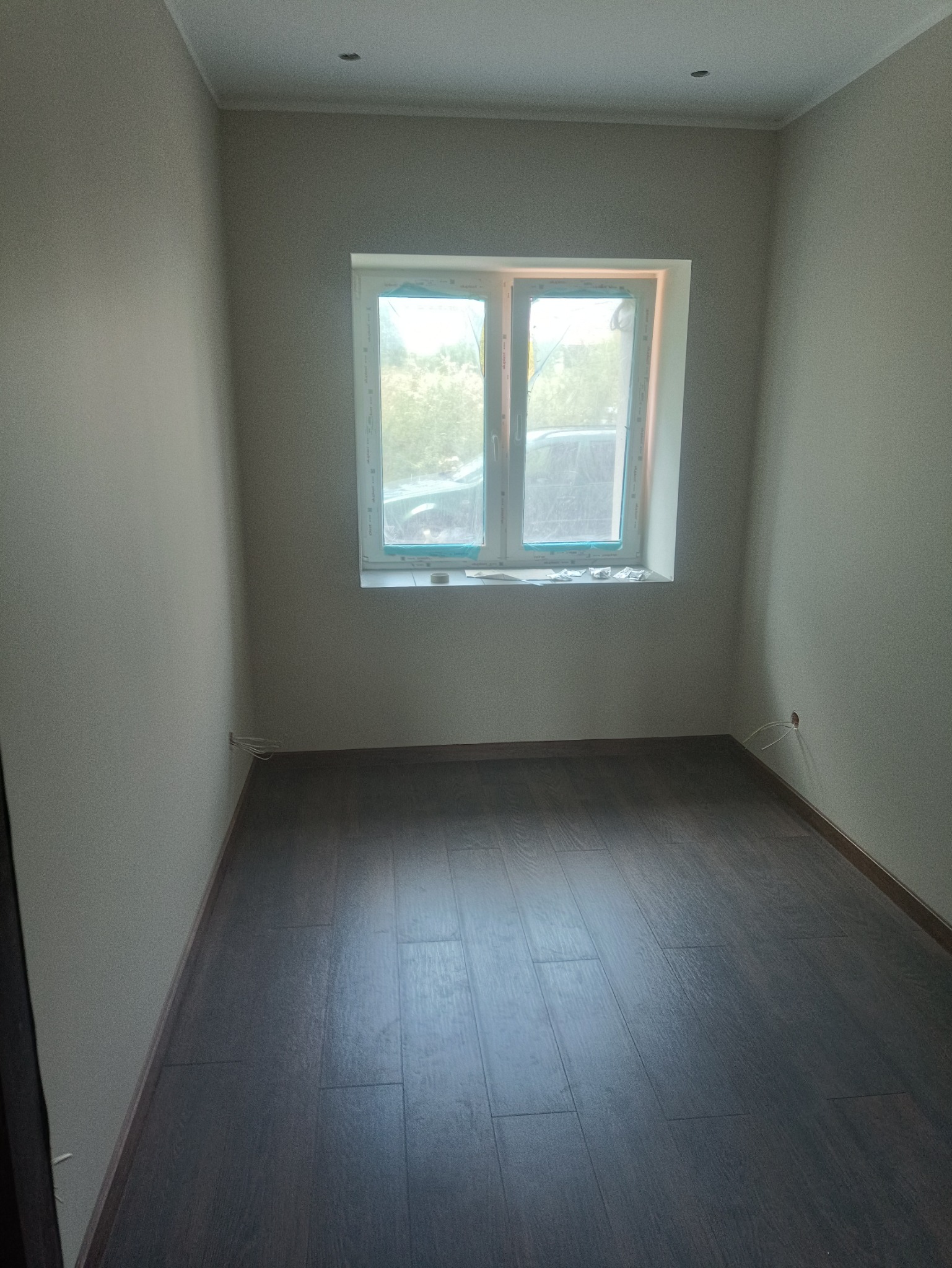 Pusty pokój po remoncie z ciemną podłogą z paneli, świeżo pomalowanymi ścianami i oknem zabezpieczonym folią malarską.