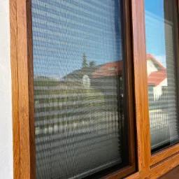 Drewniane okno z zamontowaną siatką ochronną na owady, na zewnątrz widoczne budynki i drzewa, w drugim oknie widoczne plisy.