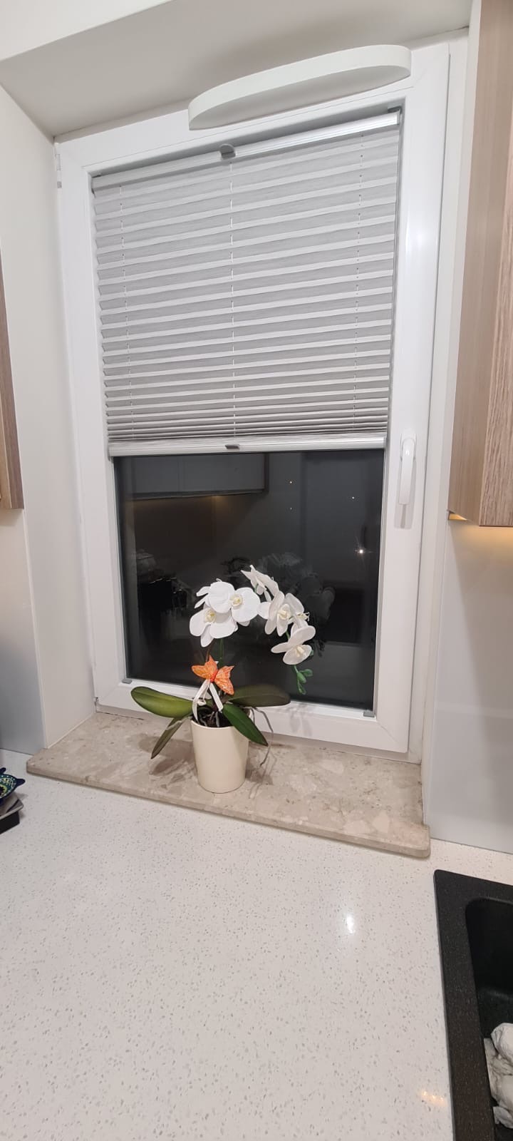 Białe okno z zamontowanymi plisami, na parapecie stoi doniczka z białą orchideą z pomarańczową ozdobą.