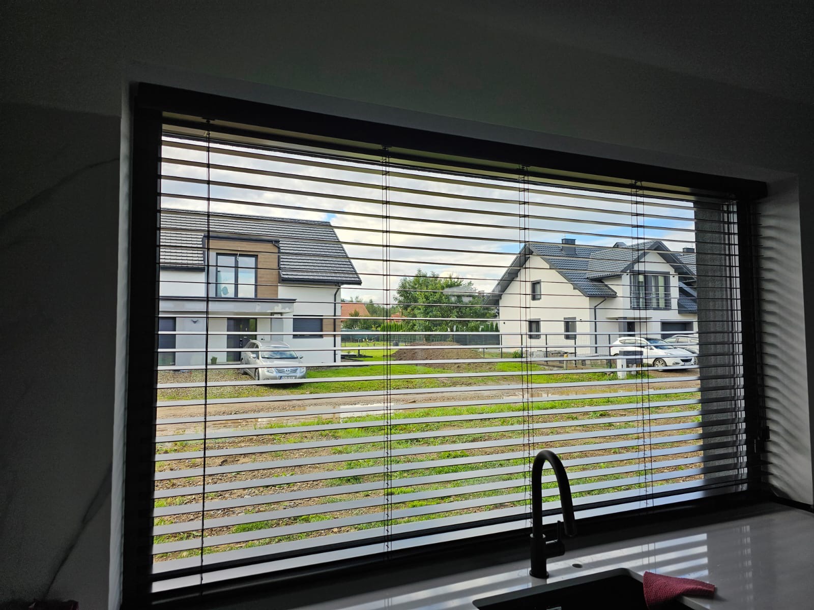 Ciemne poziome listwy osłaniają okno kuchni z widokiem na dwa nowe domy i samochody na zewnątrz.