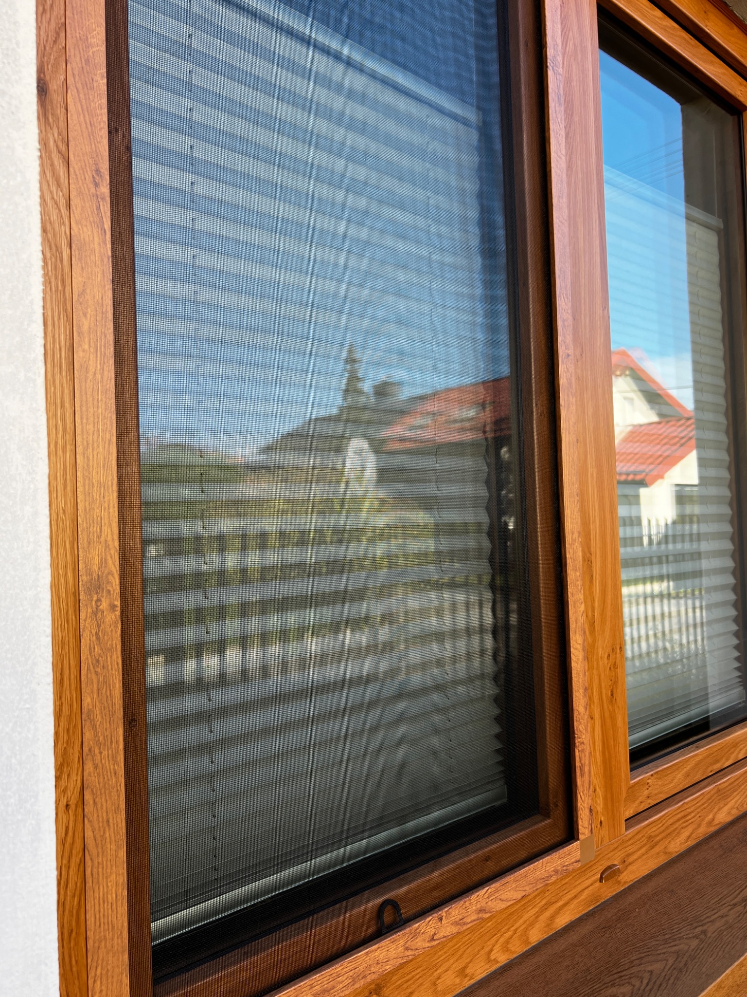 Drewniane okno z zamontowaną siatką ochronną na owady, na zewnątrz widoczne budynki i drzewa, w drugim oknie widoczne plisy.