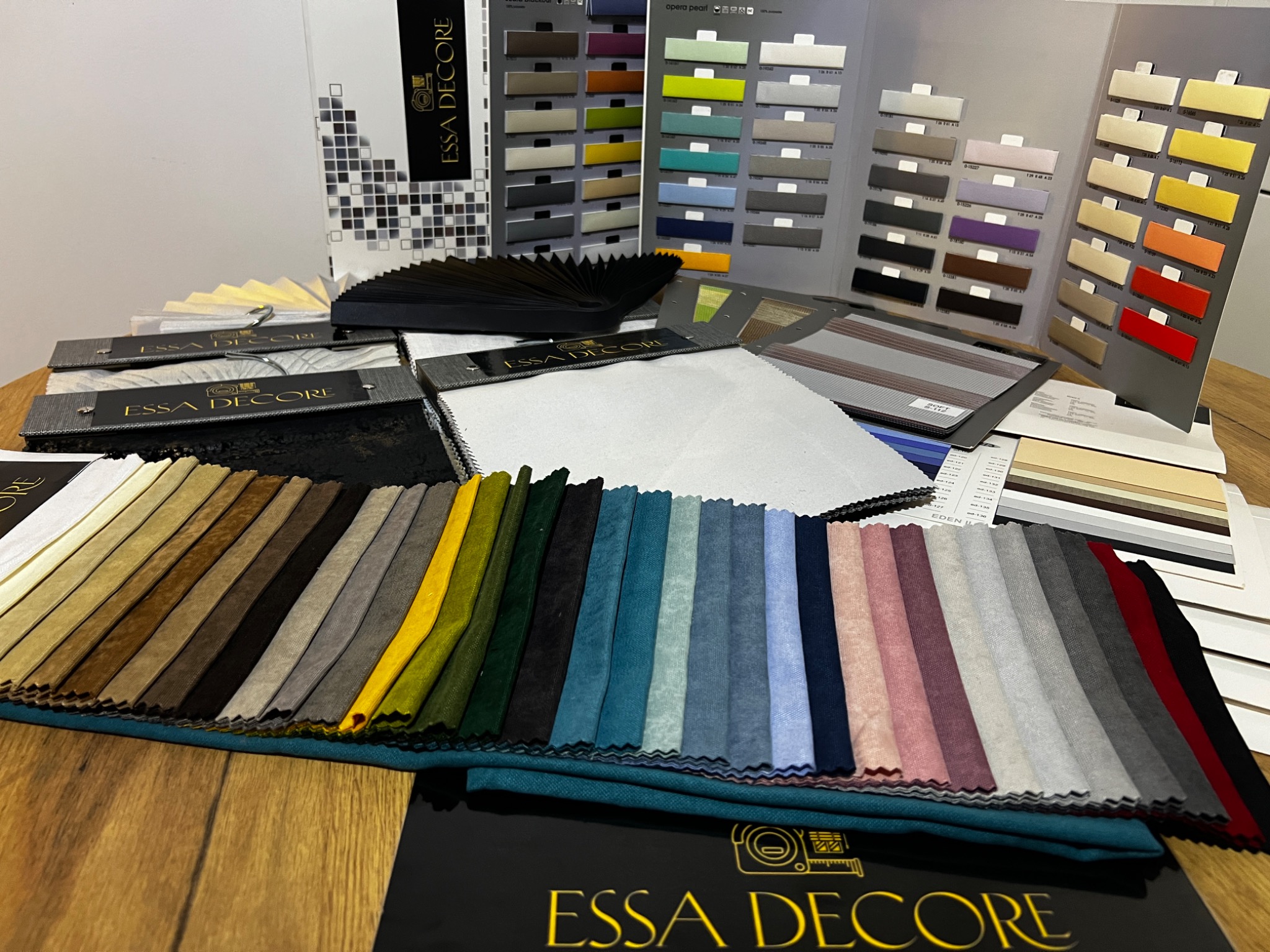 Paleta kolorów i materiałów rolet i żaluzji Essa Decore, prezentowana na drewnianym stole z katalogami i próbkami tkanin.