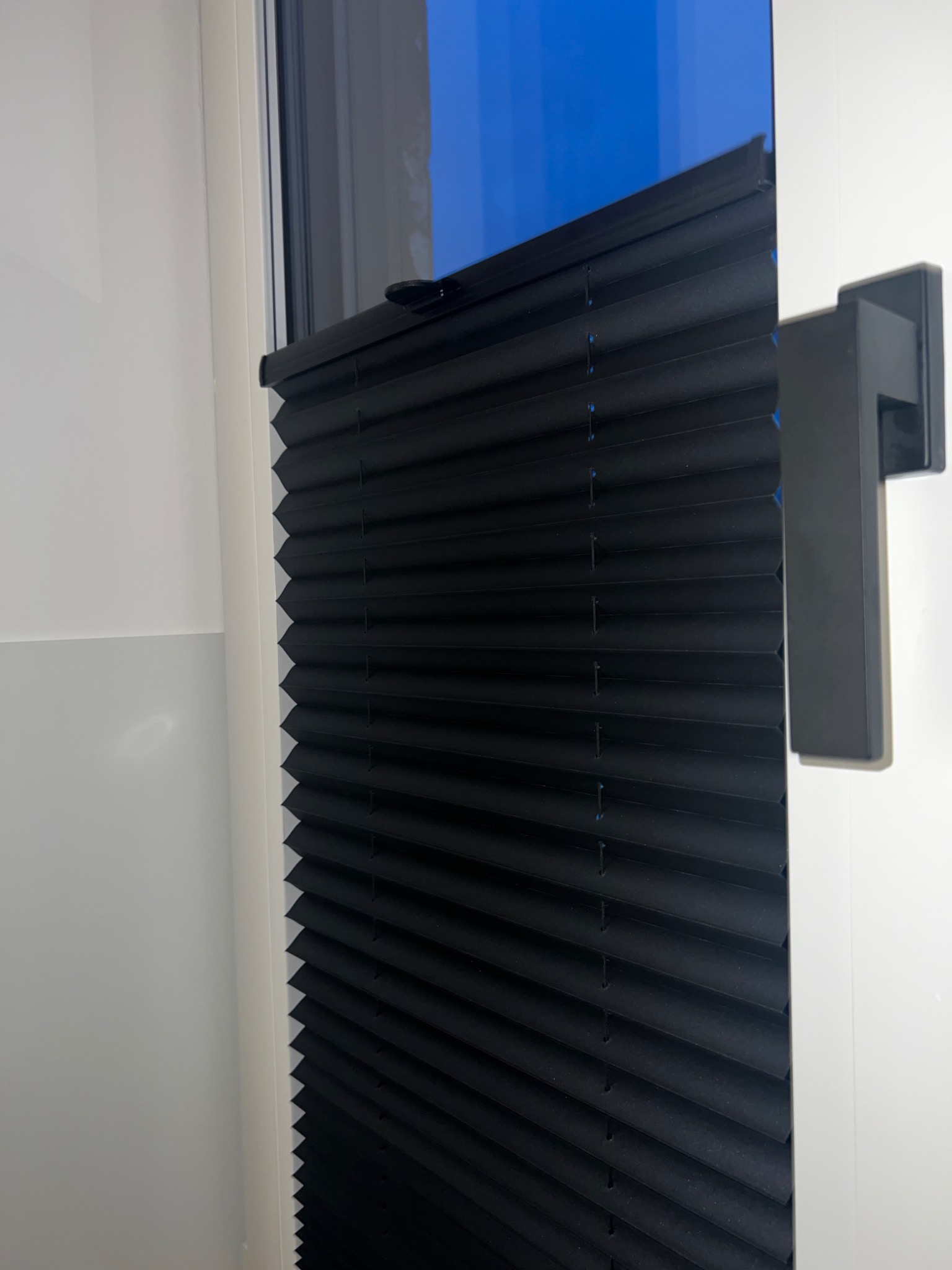 Ciemna, plisowana osłona okienna z czarnym uchwytem, zamontowana w białej ramie okiennej, widok z bliska.