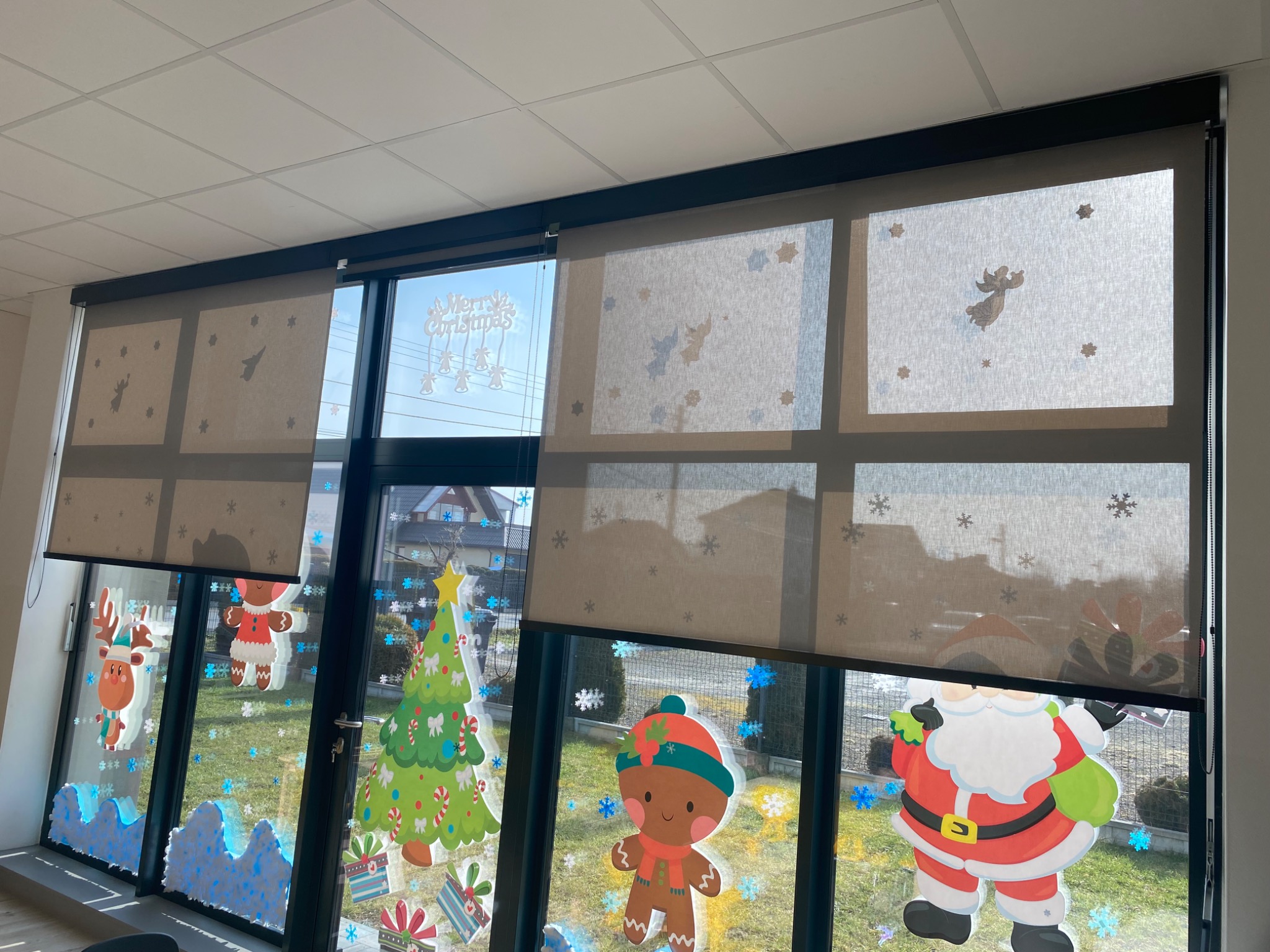 Rolety wewnętrzne w kolorze beżowym z nadrukowanymi motywami świątecznymi, częściowo zasłonięte, widoczne dekoracje okienne z motywami świątecznymi.