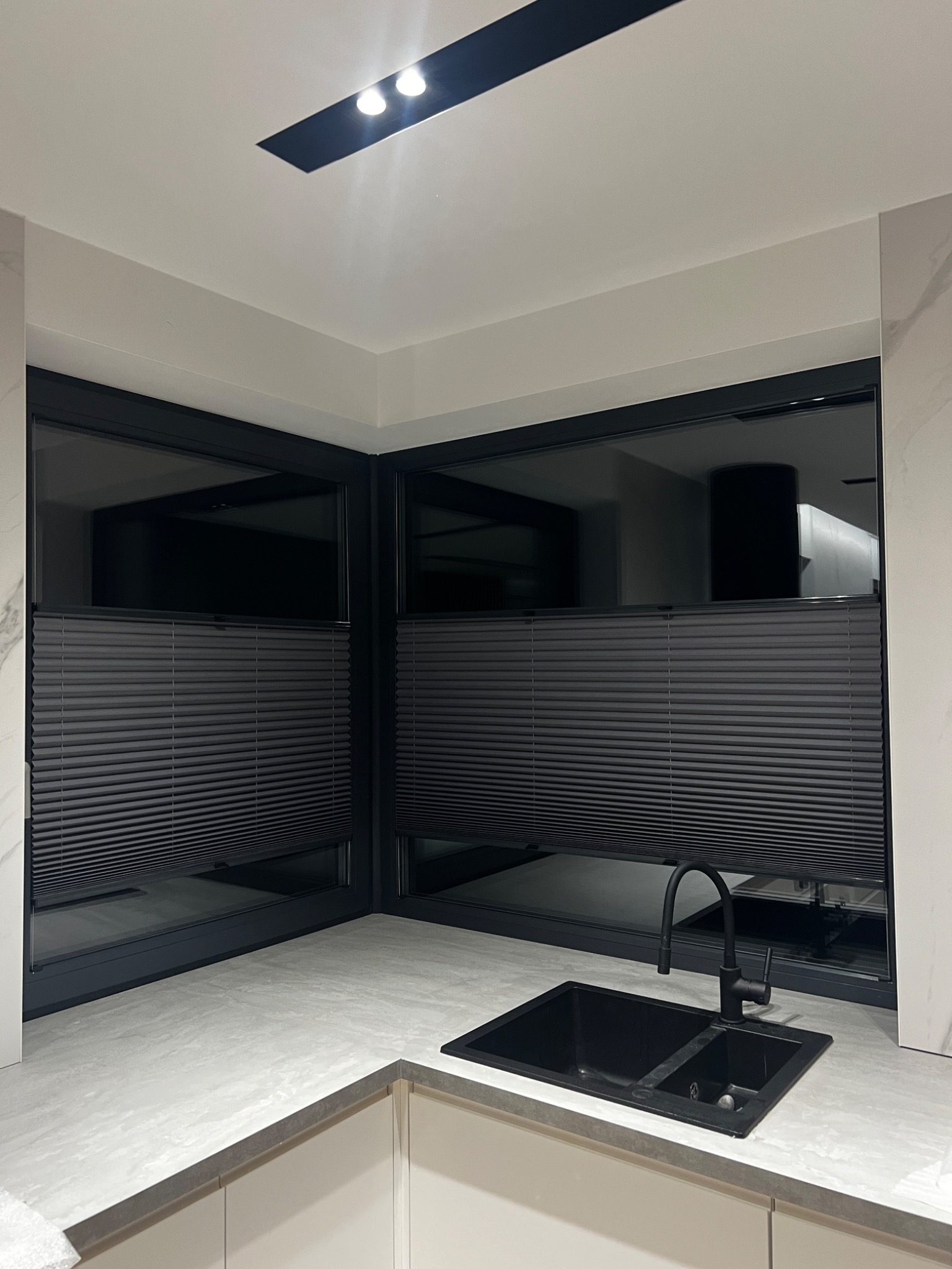 Ciemnoszare, poziome osłony okienne w kuchni z czarnym zlewem i baterią; widok na wnętrze przez okna.