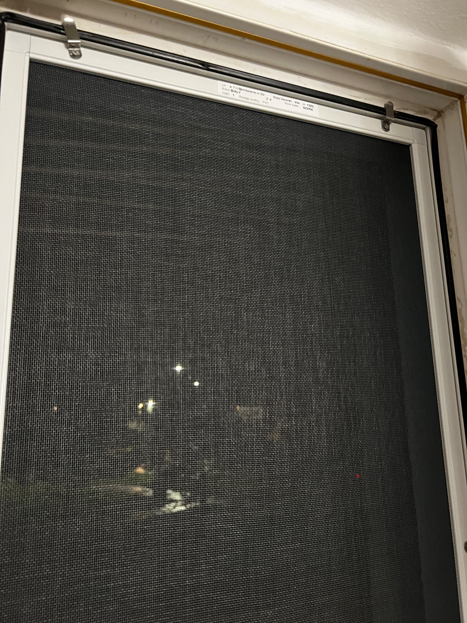 Zamknięta siatka okienna w białej ramie, widok na nocne światła miasta przez drobne oczka siatki, metalowe zaczepy mocujące u góry.