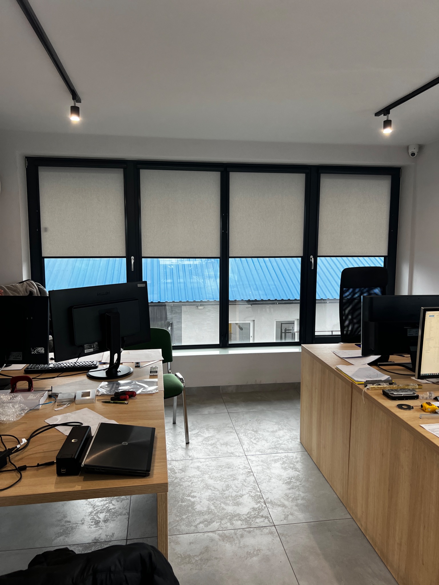 Biuro z trzema oknami z zasłoniętymi roletami w kolorze jasnego beżu, widok zza biurek z komputerami.