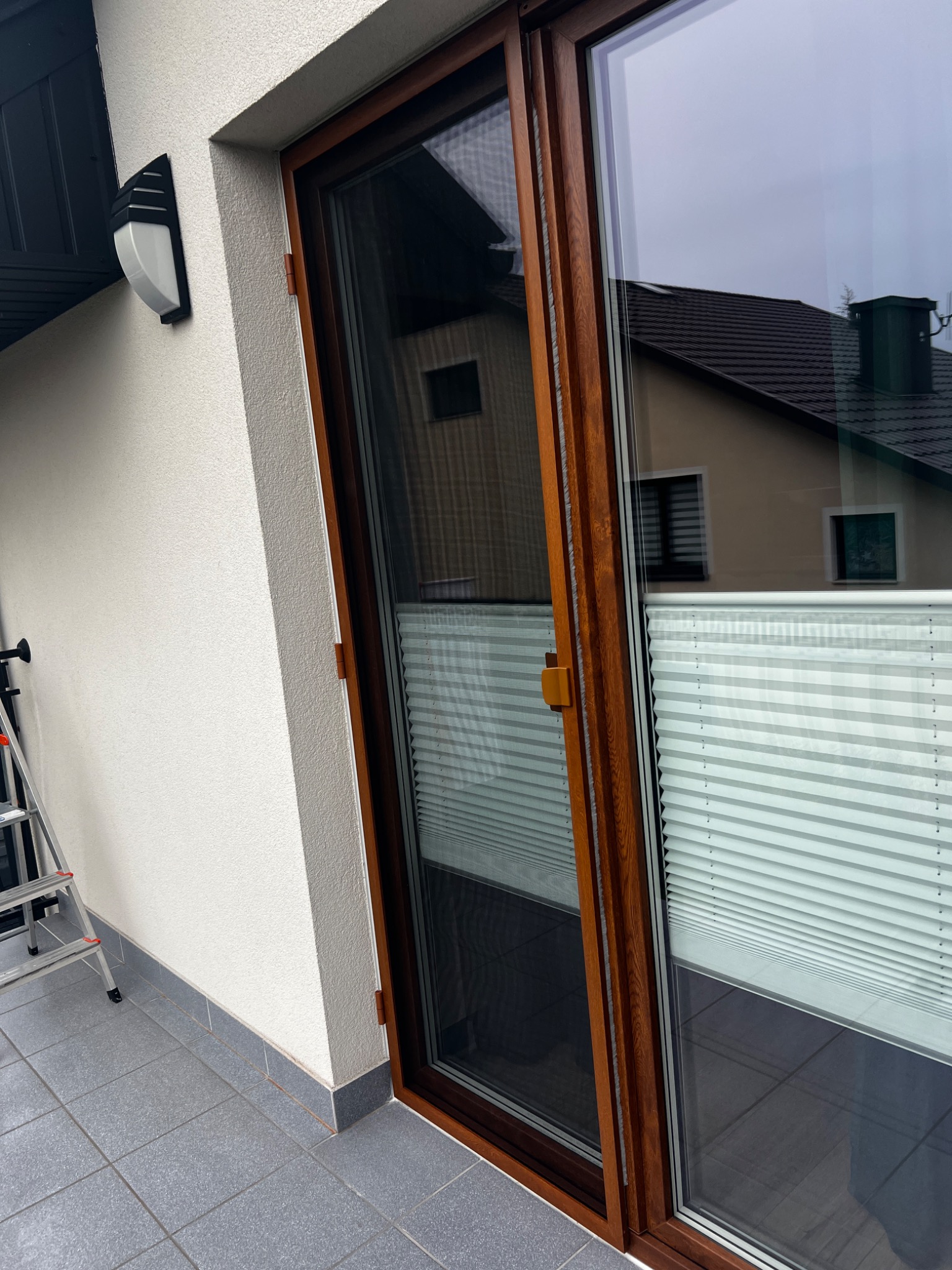 Zewnętrzne okno balkonowe z brązową ramą, wyposażone w białą plisę i ciemną moskitierę, widoczne z zewnątrz na tle elewacji domu.