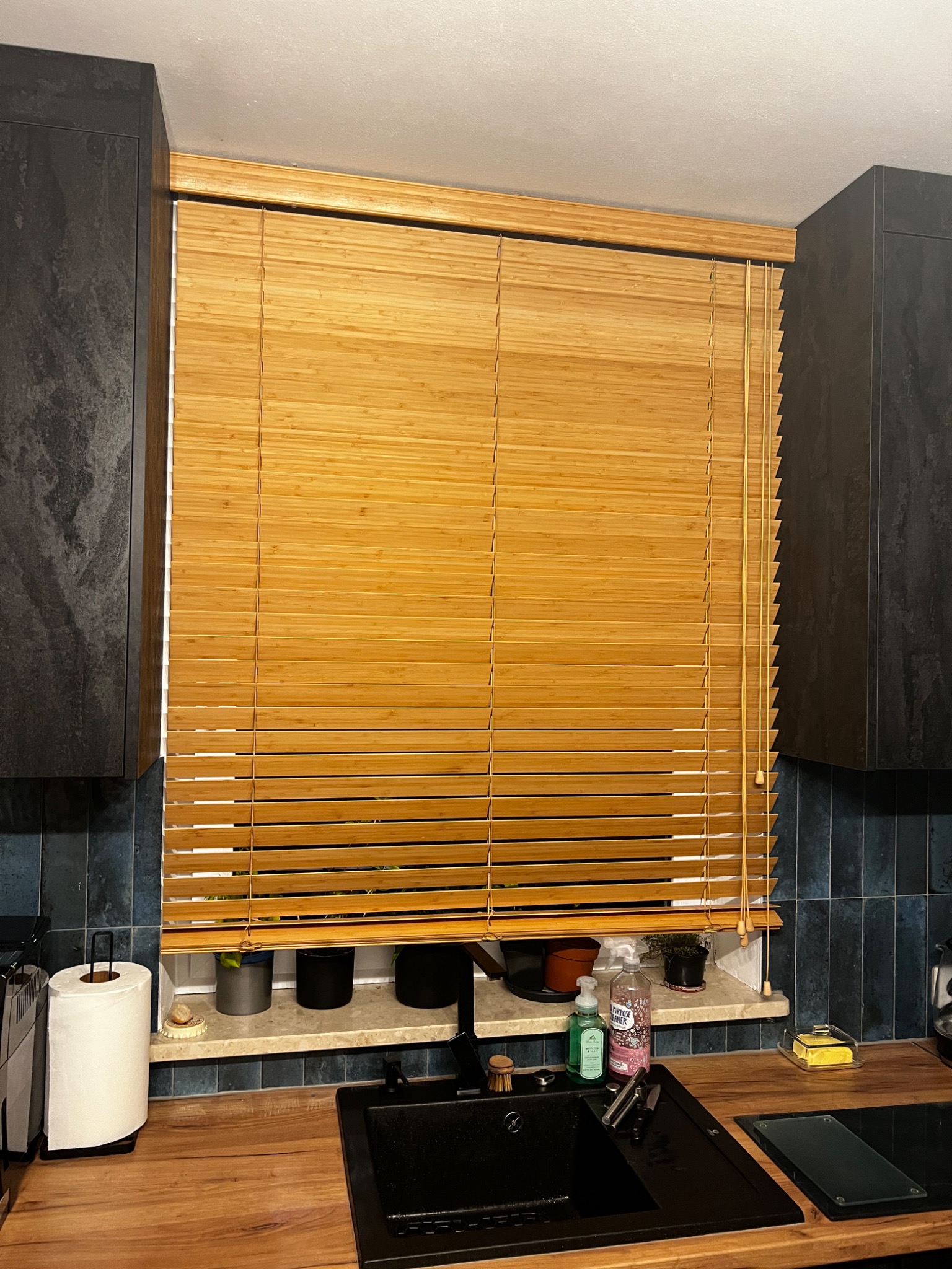 Jasne, drewniane żaluzje poziome w kuchni z ciemnymi szafkami i niebieskimi płytkami, widoczne rośliny na parapecie i akcesoria kuchenne na blacie.