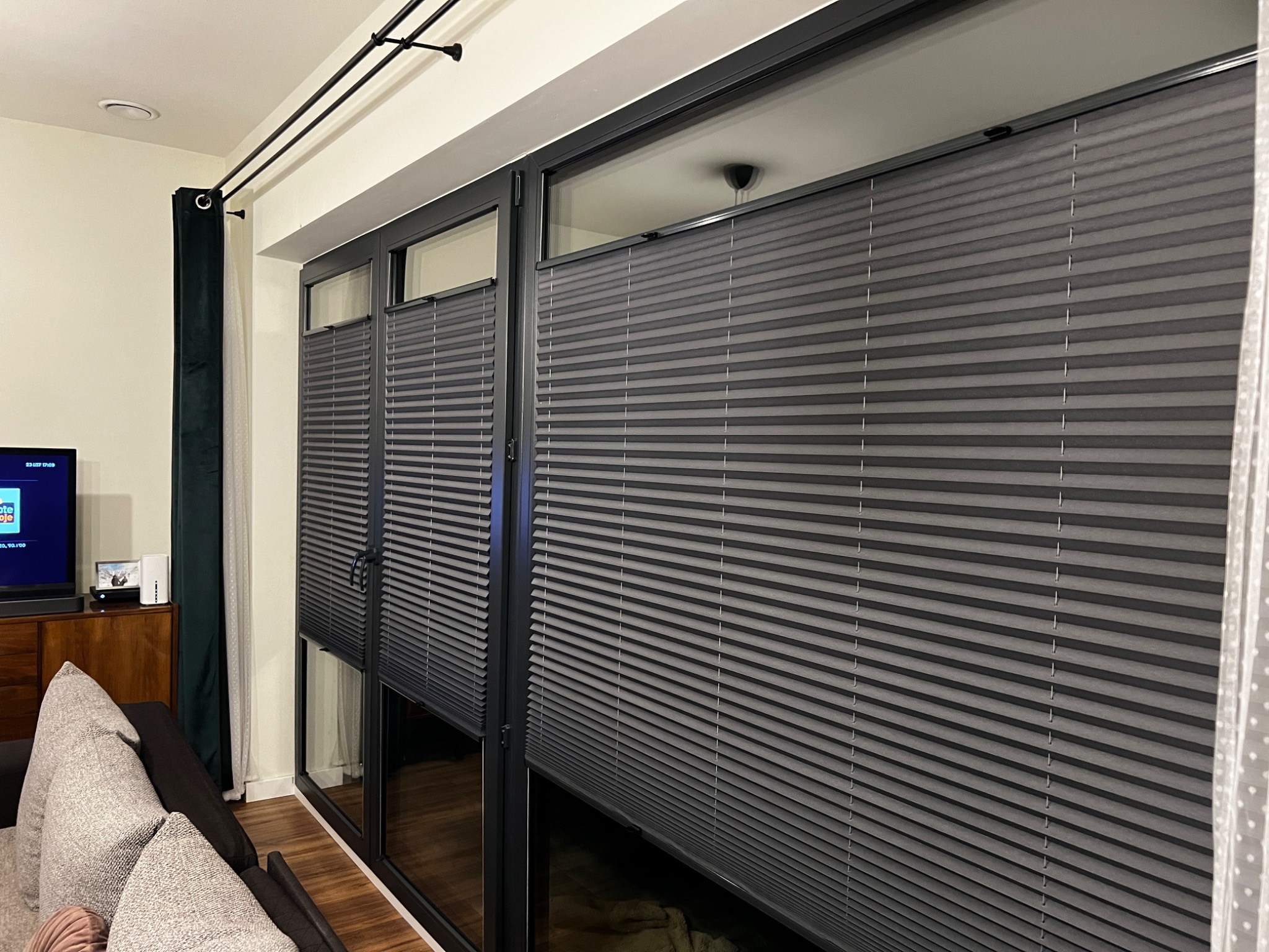 Szare plisy okienne zamontowane w oknach balkonowych, widok z wnętrza salonu z elementami wystroju.