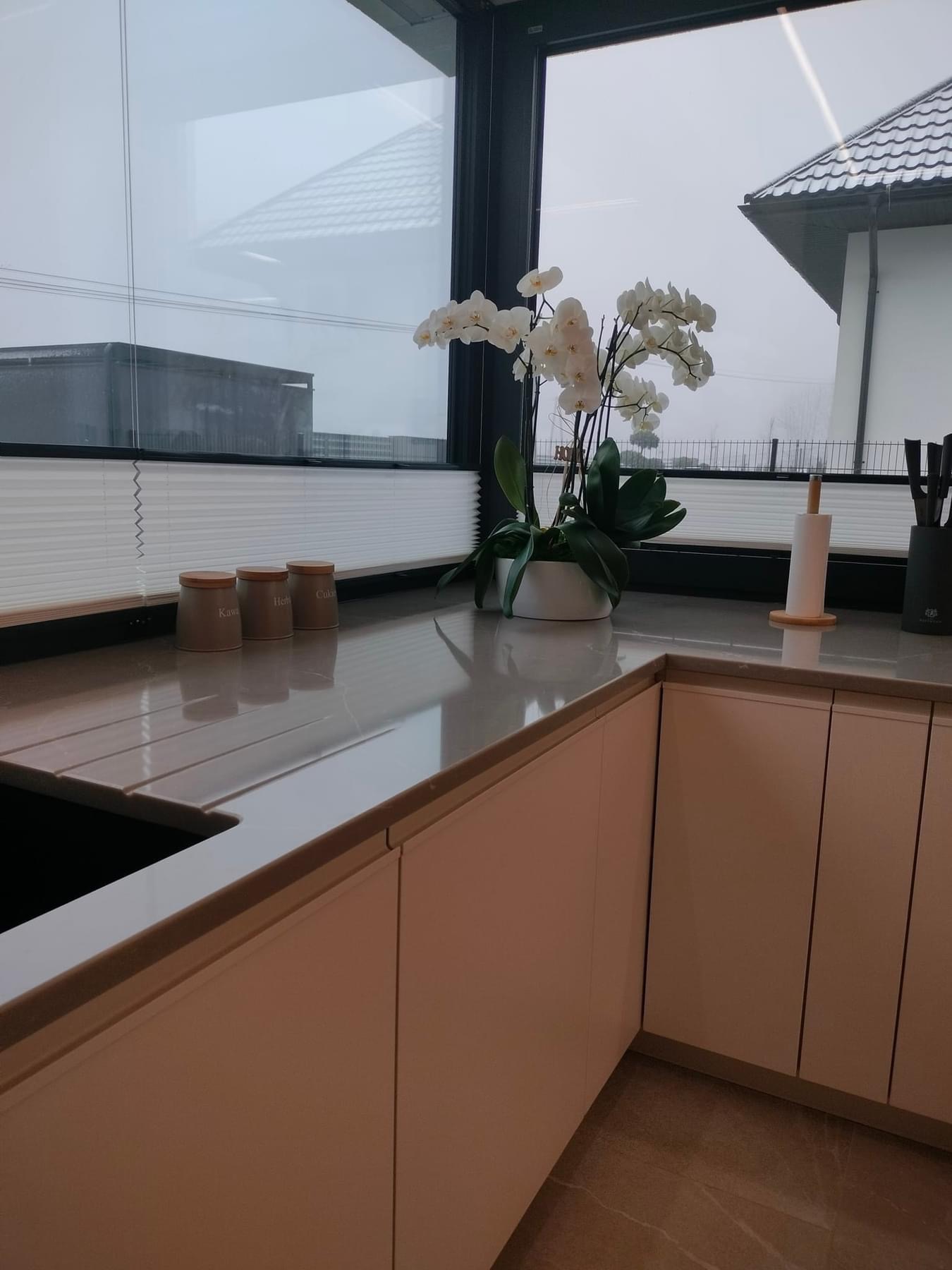 Nowoczesna kuchnia z białymi szafkami, jasnym blatem i plisami okiennymi w tle, widok na pochmurne niebo.